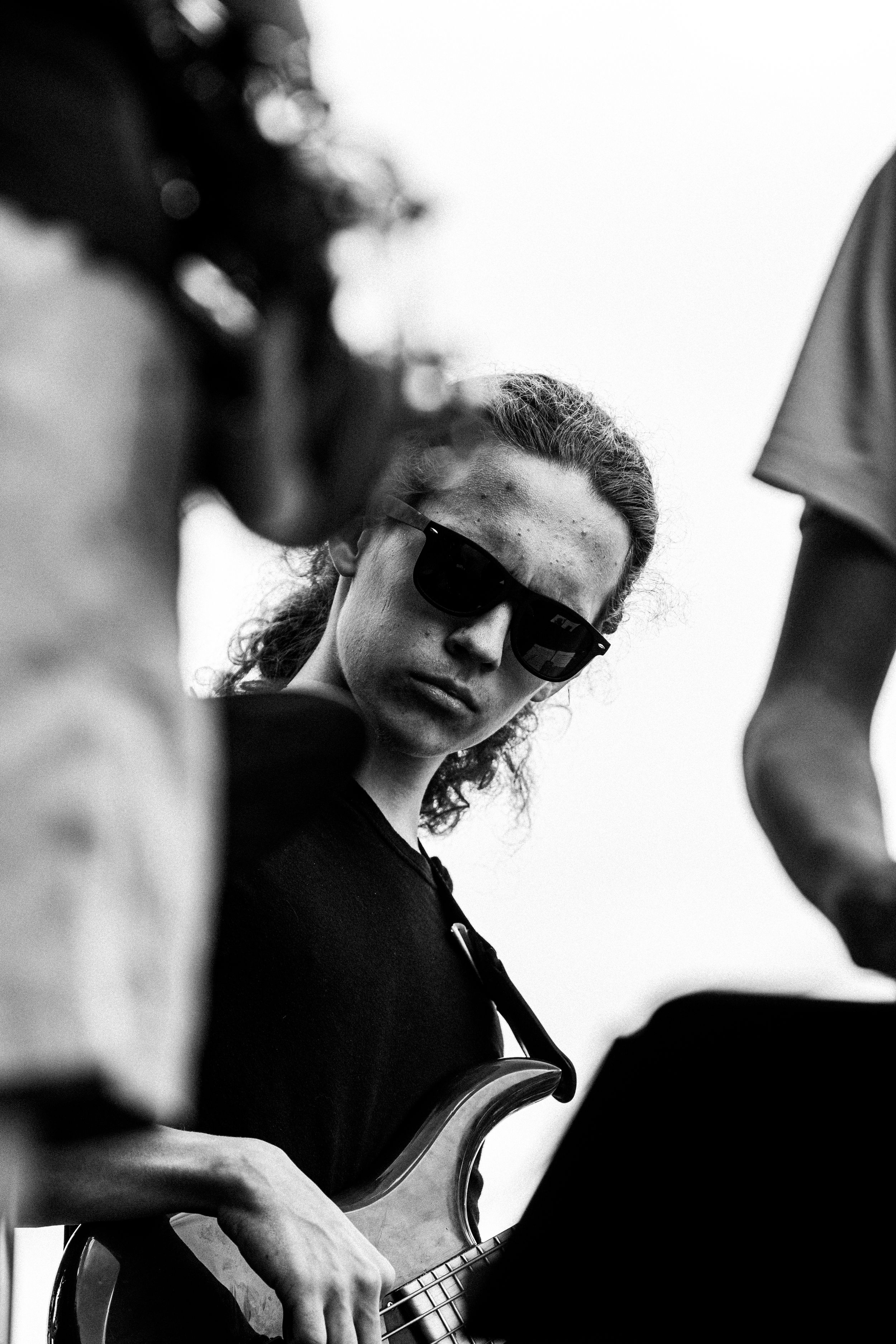 Black and white photo of a young musician with curly hair and sunglasses playing bass guitar at an outdoor concert.