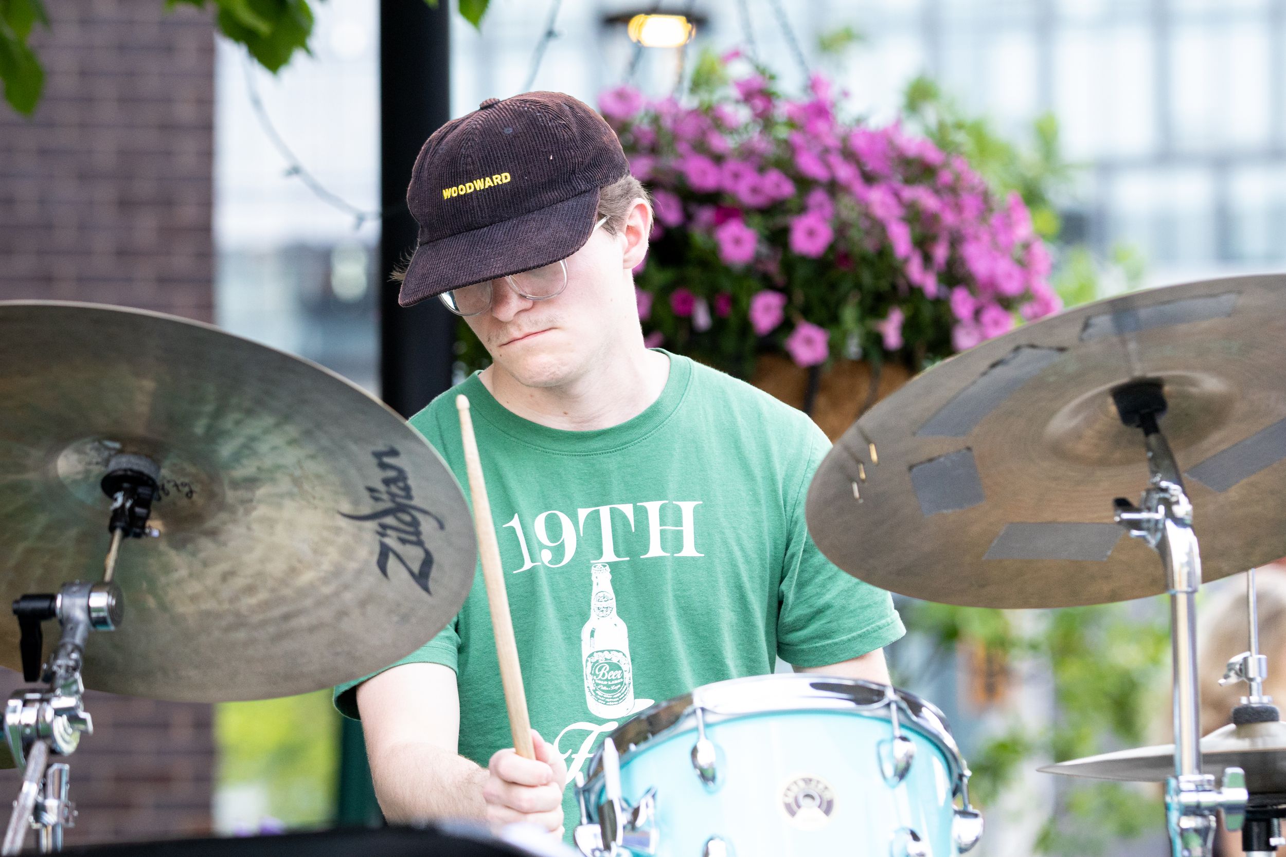 Young drummer in green 19TH t-shirt and black cap playing cymbals and snare drum outdoors.