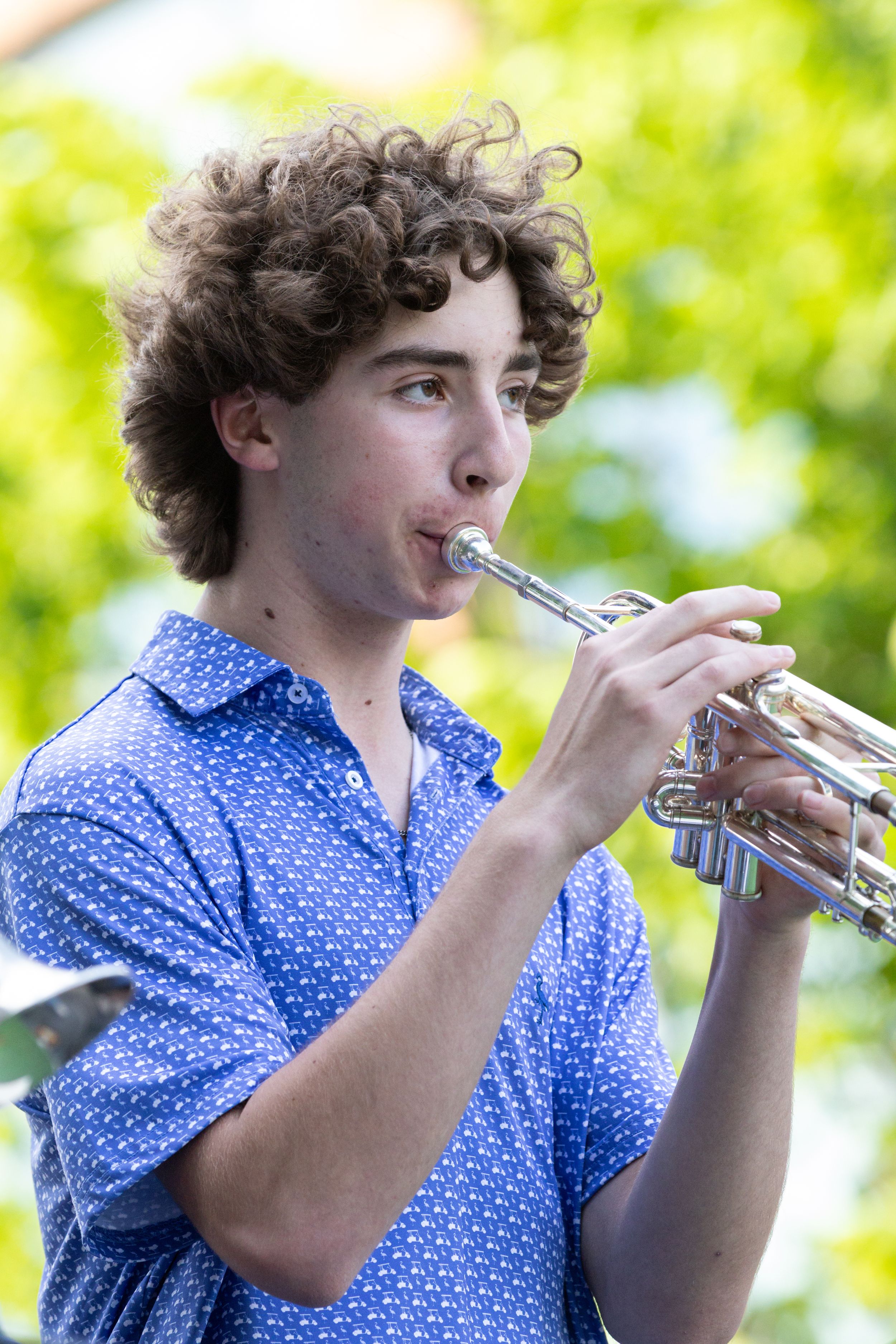 Young curly-haired musician plays trumpet outdoors in blue polka dot shirt with green trees in background.