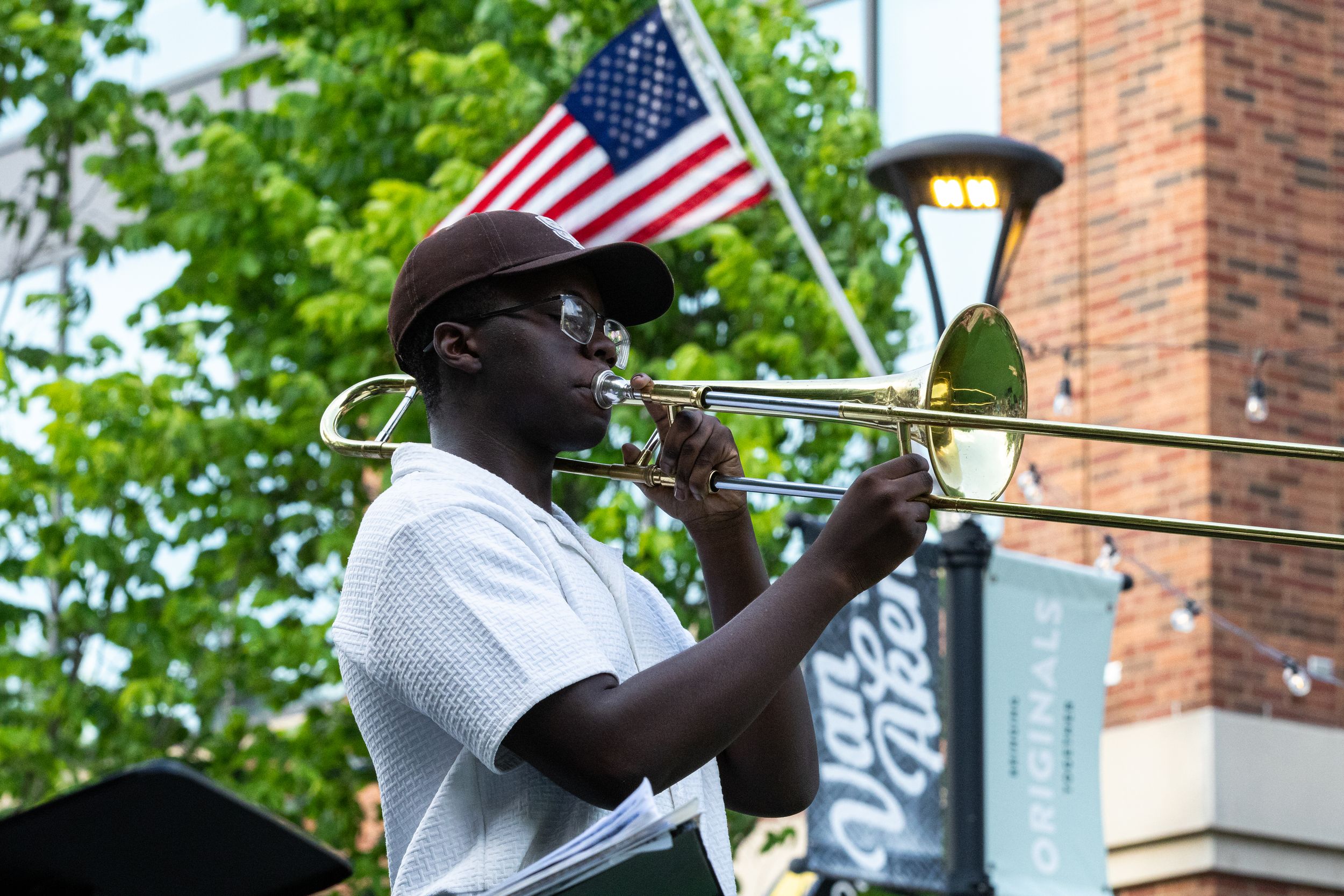 Man playing trombone outdoors at a street festival with an American flag in the background.