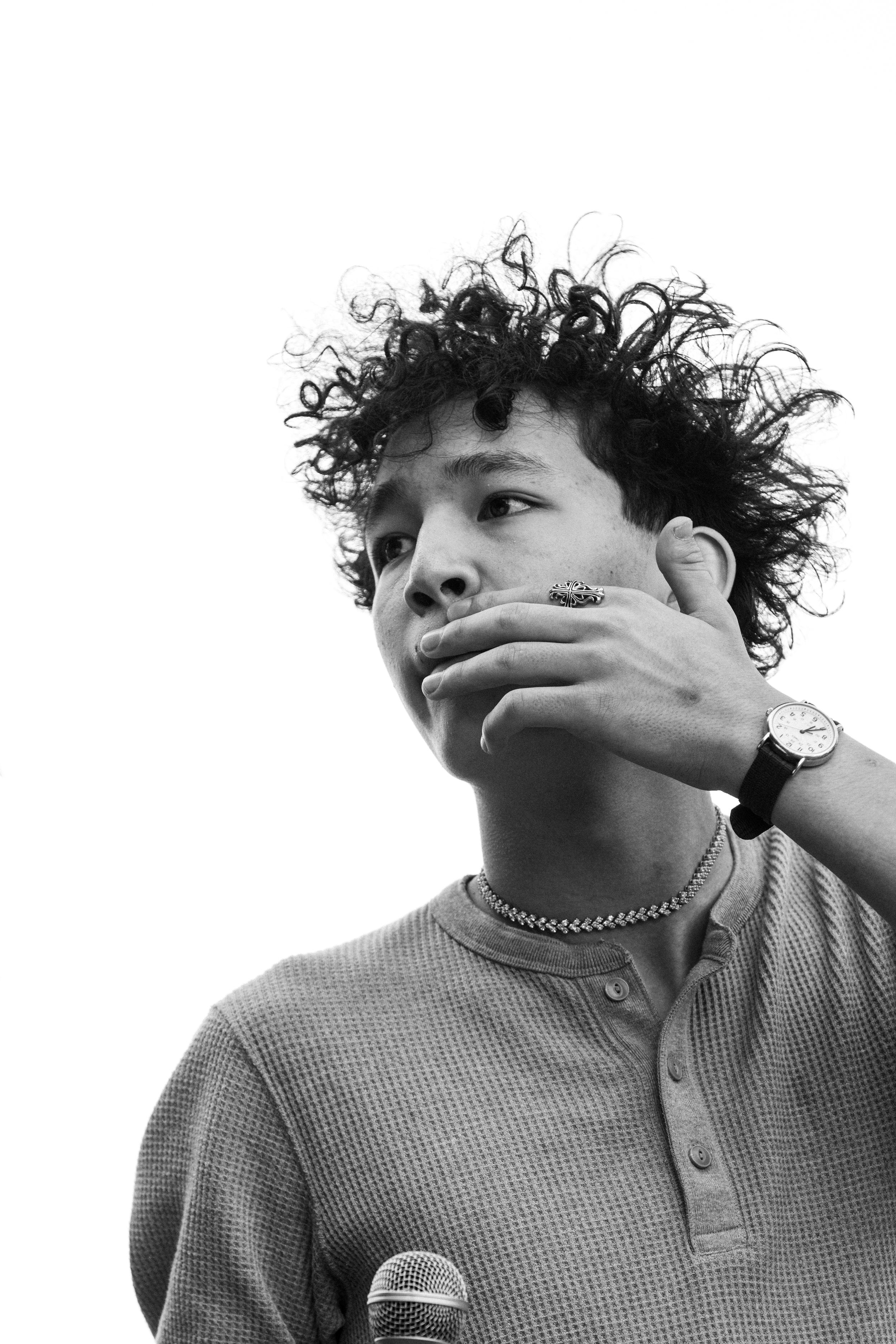 Black and white portrait of a young man with curly hair holding a microphone, wearing a watch and ring.