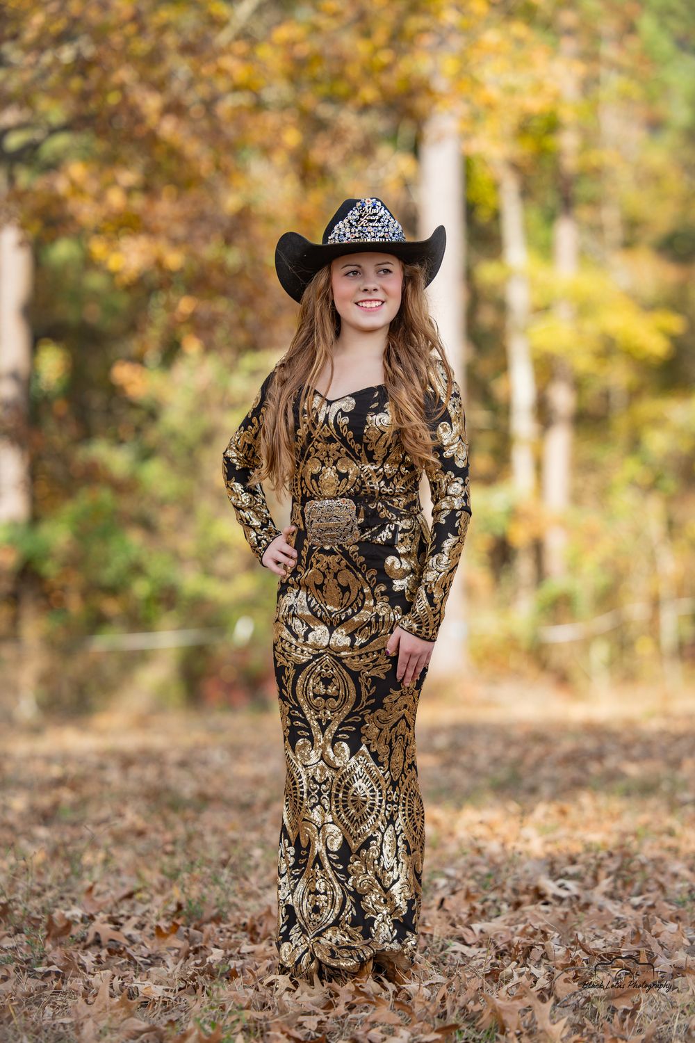 Young woman in gold and black patterned gown and black cowboy hat poses outdoors among autumn foliage.