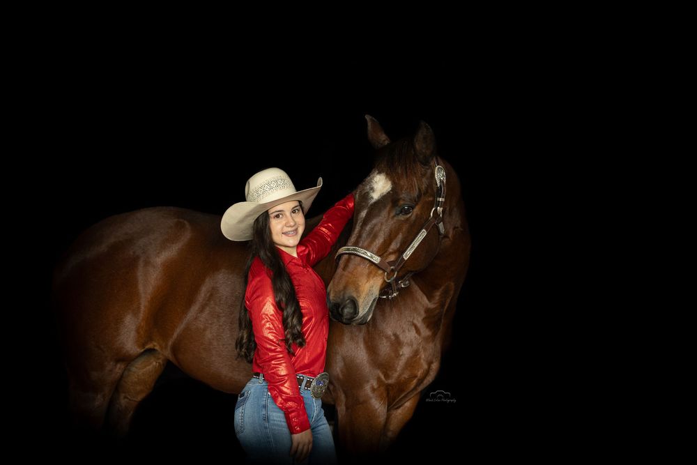 A young woman in a red jacket and white cowboy hat poses with a chestnut horse against a black background.
