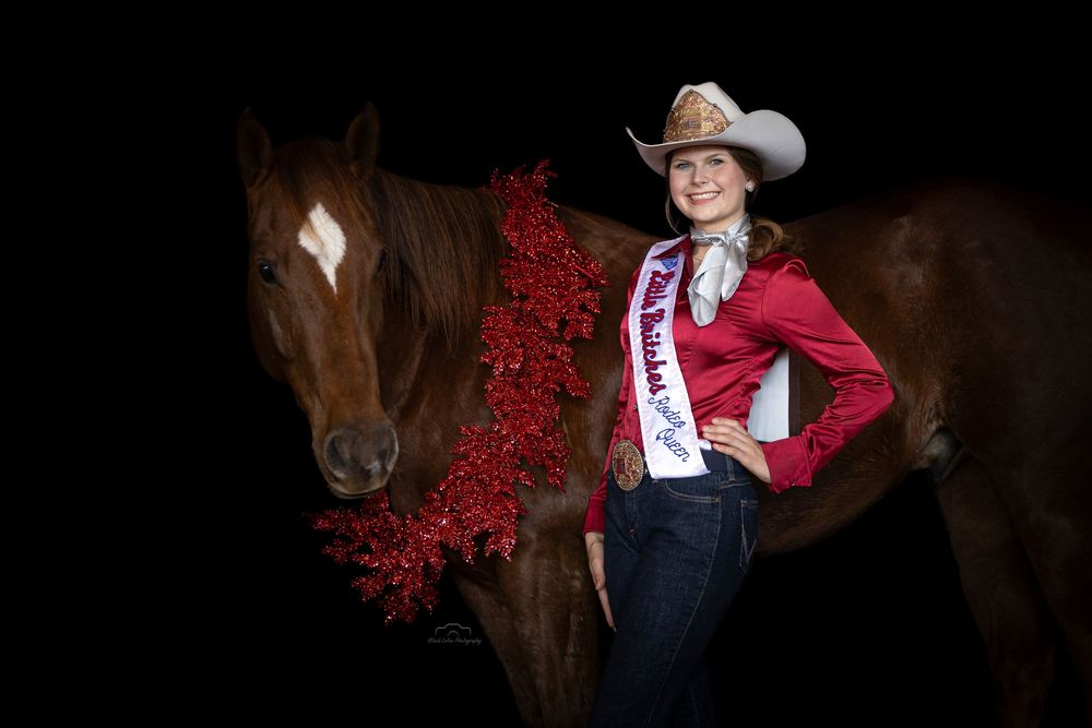 Young woman in cowboy hat and red sash poses beside a chestnut horse adorned with red tinsel garland.