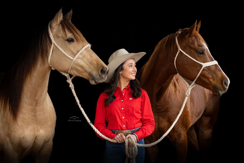 Woman in red shirt and cowboy hat stands between two horses on black background.