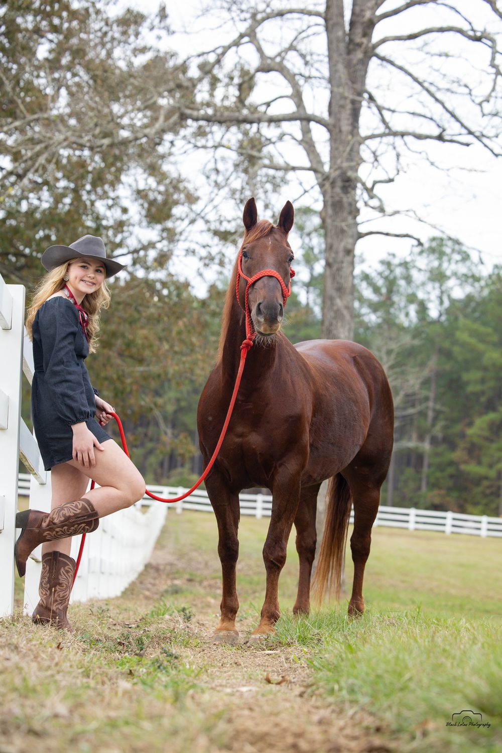 Young woman in cowboy hat and boots poses with a chestnut horse on a red lead rope at a white fence ranch.
