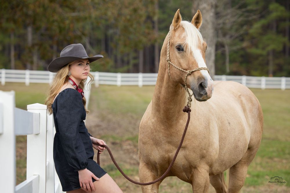 A blonde cowgirl in a black hat and jacket poses with a palomino horse at a white fence ranch.
