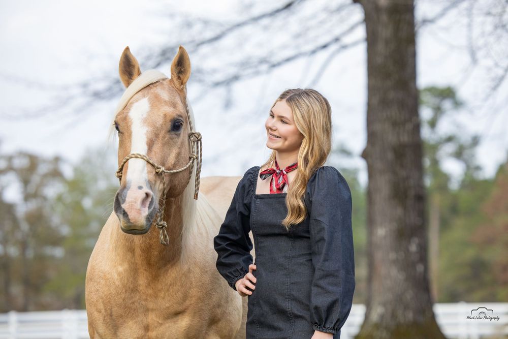 Blonde woman in black jacket stands beside a palomino horse outdoors near trees.