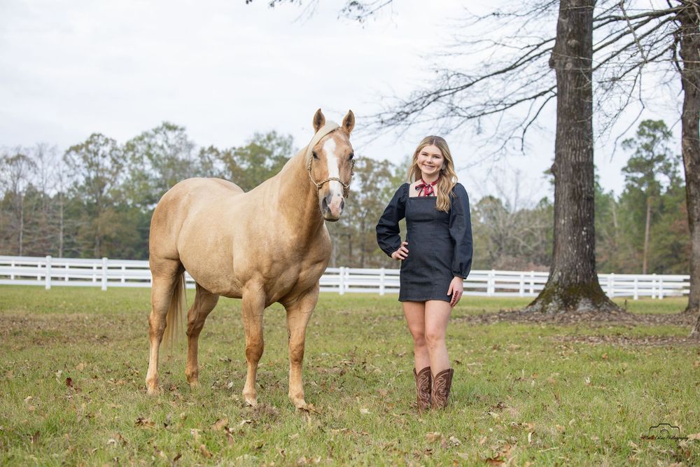 Young woman in black dress and cowboy boots stands beside a palomino horse in a green pasture with white fence.