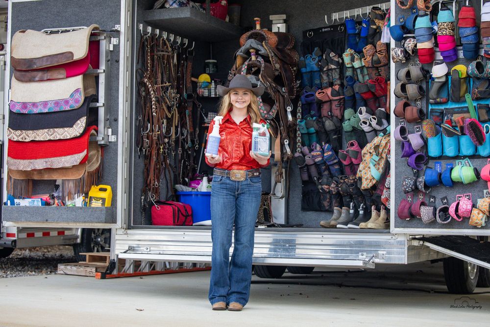 Woman in western attire stands proudly in front of a horse trailer stocked with colorful equestrian supplies and tack.