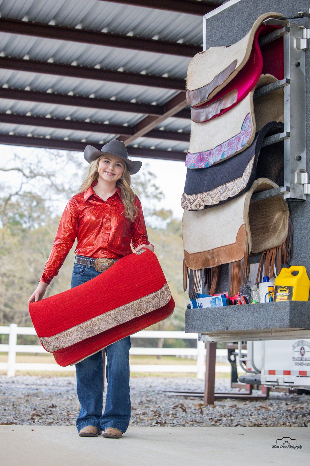 Young woman in red shirt and cowboy hat carrying a red saddle blanket near a horse trailer with saddles.