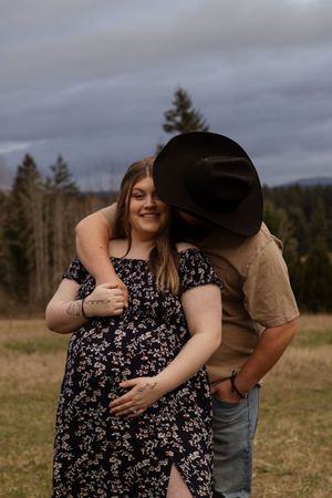 Pregnant woman in floral dress embraced by man in cowboy hat in a field in pierce county washington taken by local photographer