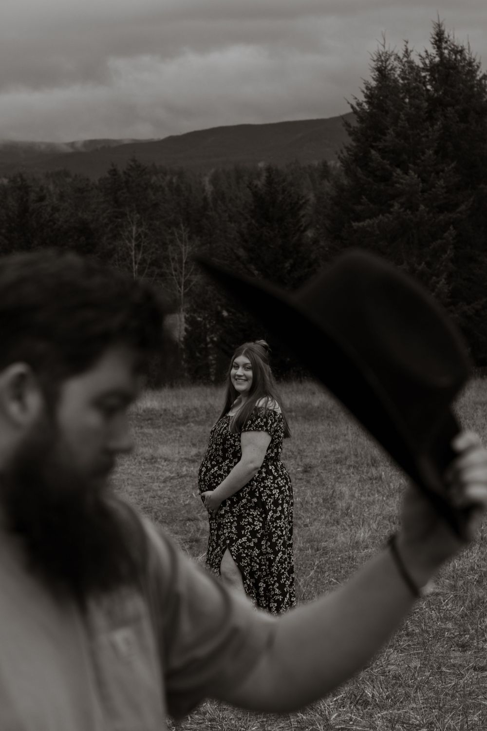 Black and white photo of a pregnant woman smiling in a field while a bearded man holds a cowboy hat taken by pierce county photographer