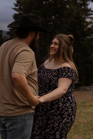 Smiling pregnant woman holding hands with bearded man in cowboy hat in eatonville washington field taken by pierce county photographer