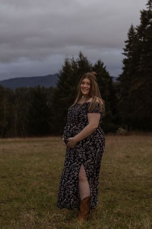 Smiling pregnant woman in floral dress standing in a field with mountains behind in Eatonville Washington near JBLM taken by WA photographer