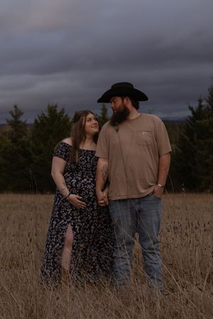 Pregnant couple holding hands in golden field at dusk with evergreen trees in eatonville washington taken by pierce county photographer
