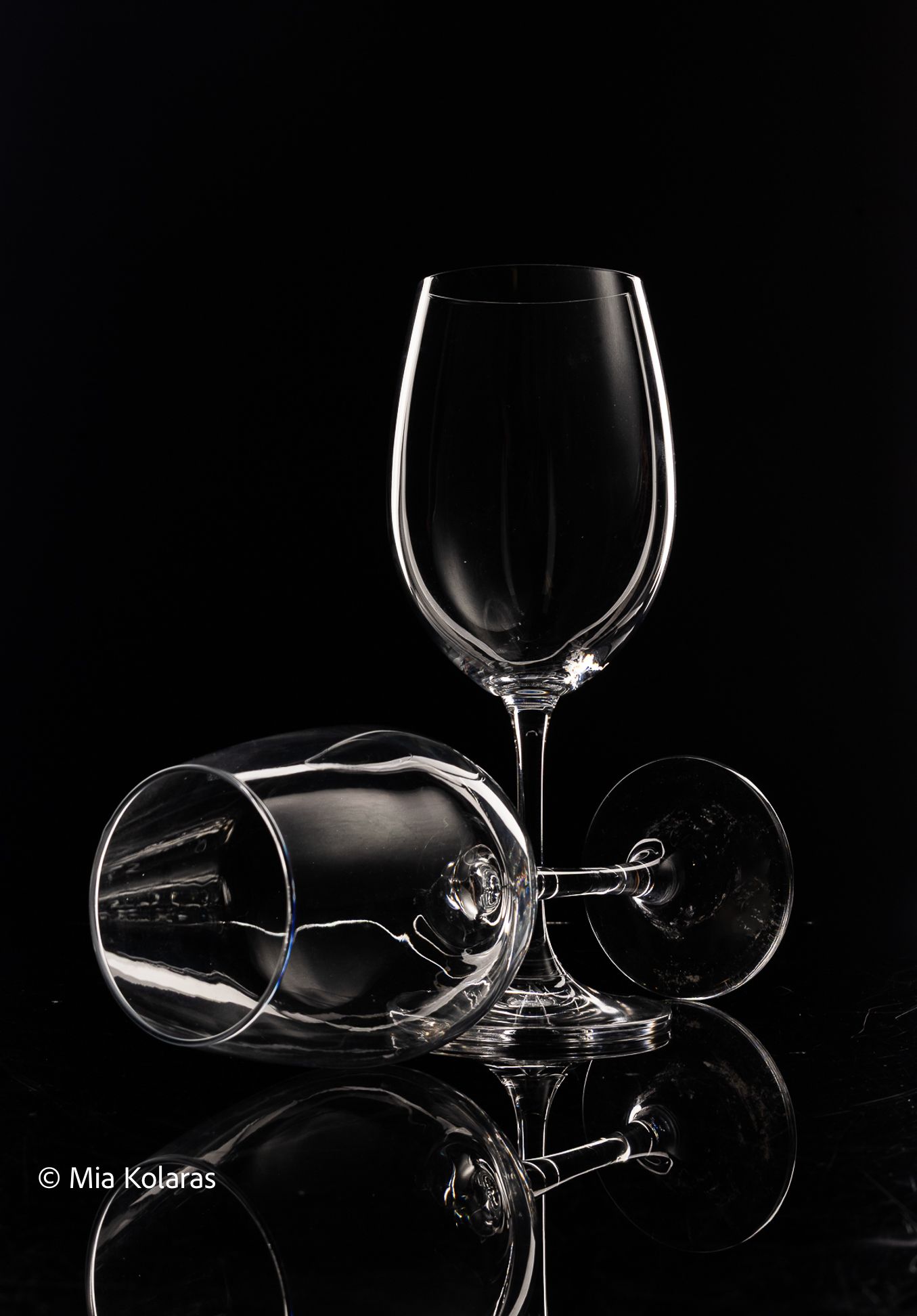 White line glass photo of two wine glasses with one standing tall and one lying down, against a black backdrop & reflective surface