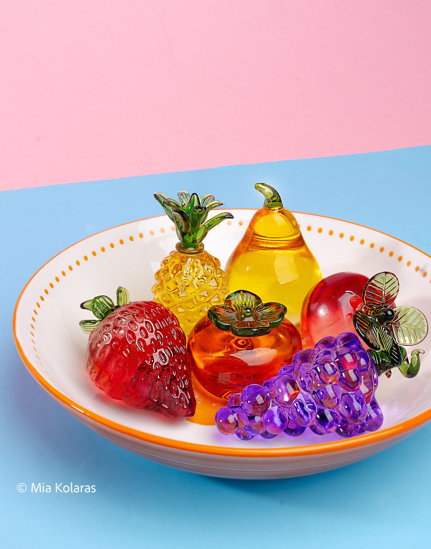 Glass fruits in a orange and white bowl, against and light blue and pink backdrop
