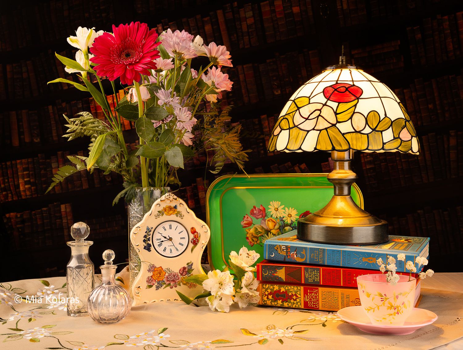 Eclectic inspired still life with a stained glass lamp, flower bouquet, glass perfume bottles, floral clock, books, tray and pink teacup