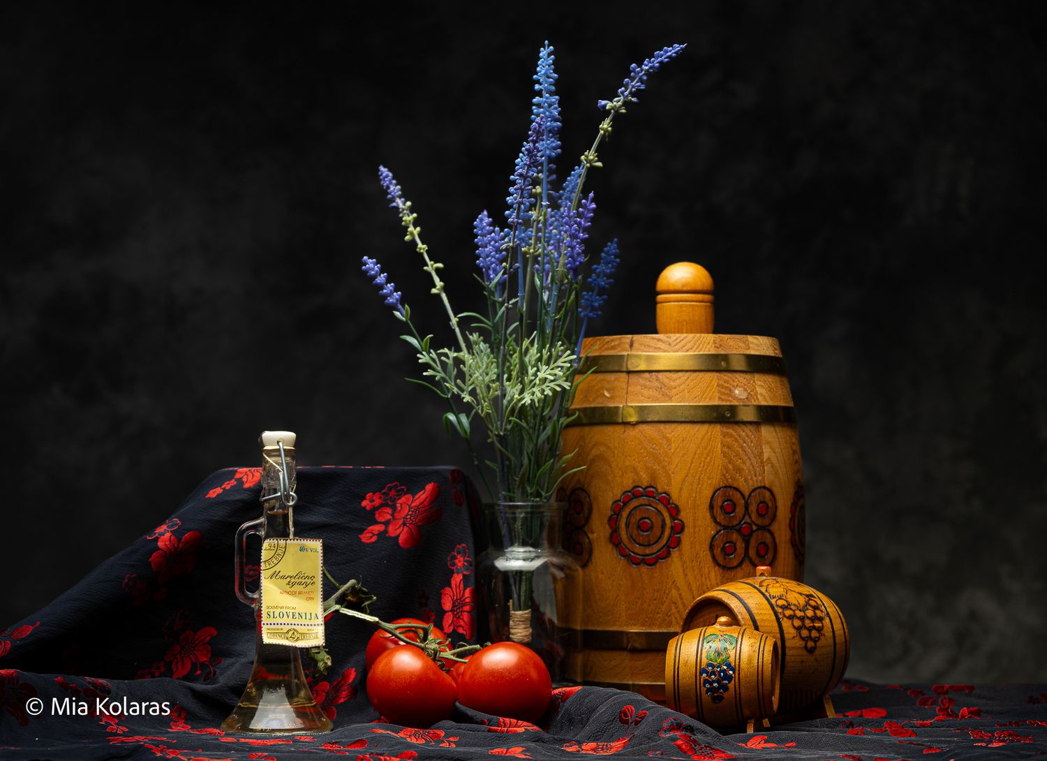 Slovenian-inspired objects in front of a grey backdrop with tomatoes, a wine glass, purple flower & alcohol