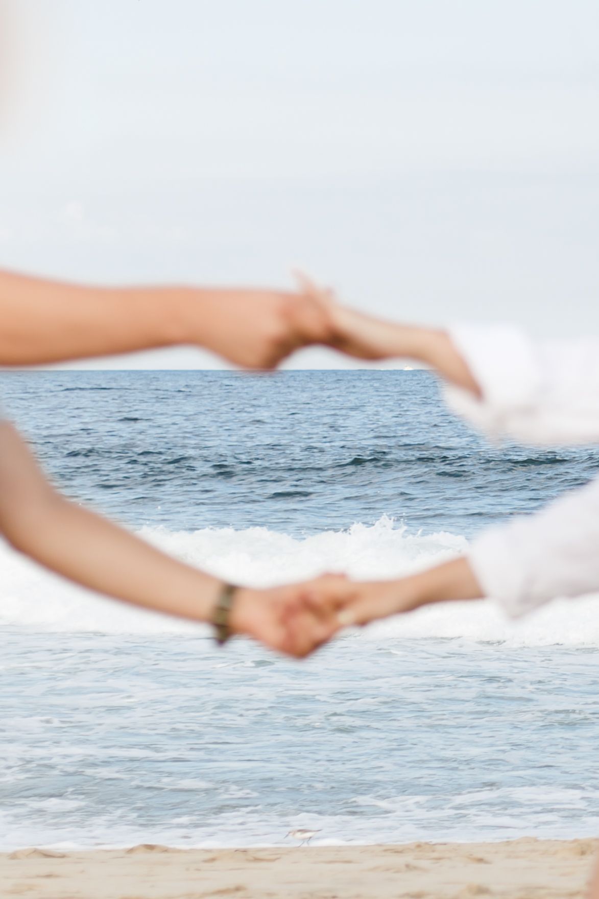 Two hands form a heart shape against an ocean backdrop with waves rolling onto a sandy beach.