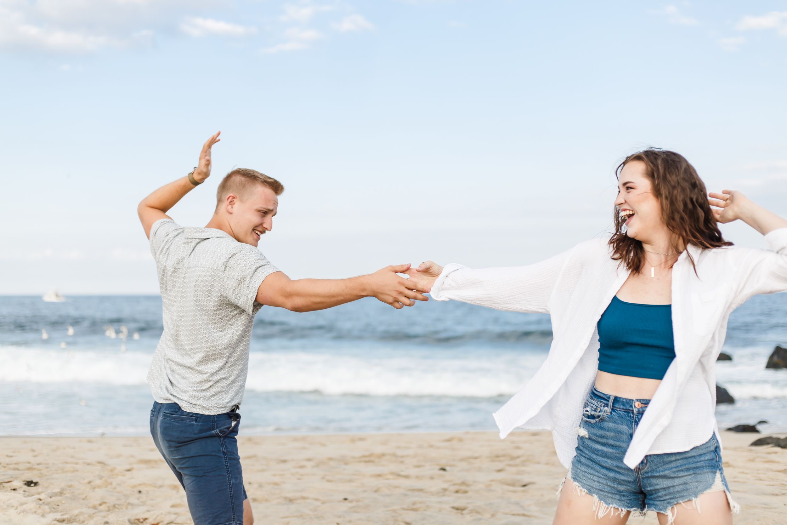 A couple dances together playfully on a scenic beach with ocean waves and mountains in the background.