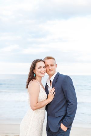 A couple poses for wedding photos on a sunny beach with ocean waves behind them.