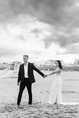 A couple in wedding attire poses playfully on a sandy beach with beach houses in the background.