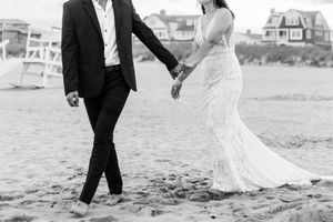 A couple in formal attire walks along a sandy beach with houses in the background.