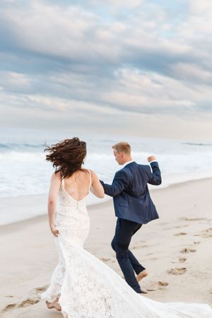 A couple runs along an empty beach with dramatic cloudy skies as her white lace dress flows in the wind.