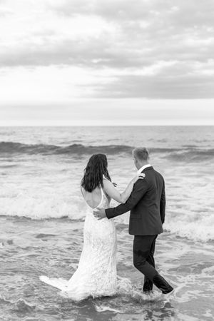 A couple in formal wear splashes through ocean waves during a beach photoshoot.