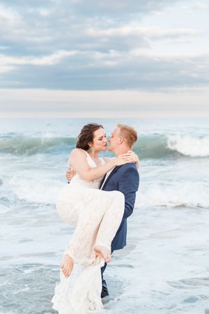 A romantic couple embraces in wedding attire as waves crash behind them on the beach.