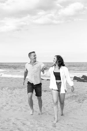 A couple walks hand in hand along a sandy beach with ocean waves in the background.