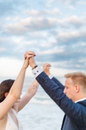 Two people hold hands raised against a cloudy beach sky in a joyful celebratory pose.