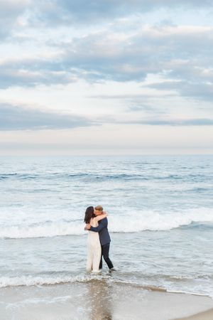 A romantic embrace between newlyweds on the shoreline with gentle waves and soft clouds in the background.