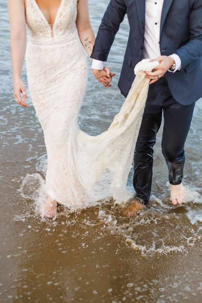 Close-up of bride's white lace dress getting wet as couple walks through ocean surf.