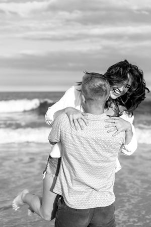 A romantic couple embraces while standing on the beach with waves crashing behind them.
