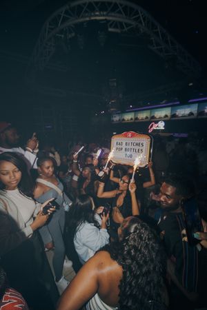 Crowded nightclub party scene with people dancing and holding a lit sign reading Bad Bitches Pop Bottles Too.