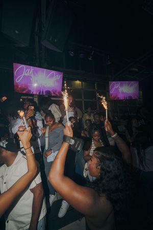 Crowd celebrating at a nightclub party with sparklers, purple screens displaying event branding in the background.