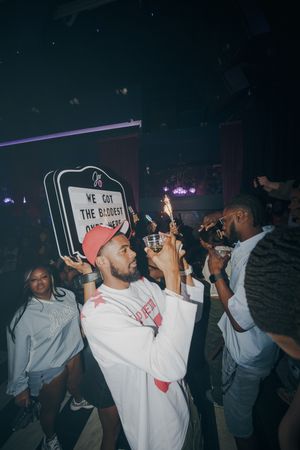 People celebrating at a nightclub party, man in white hoodie holding sparkler drinks on dance floor.