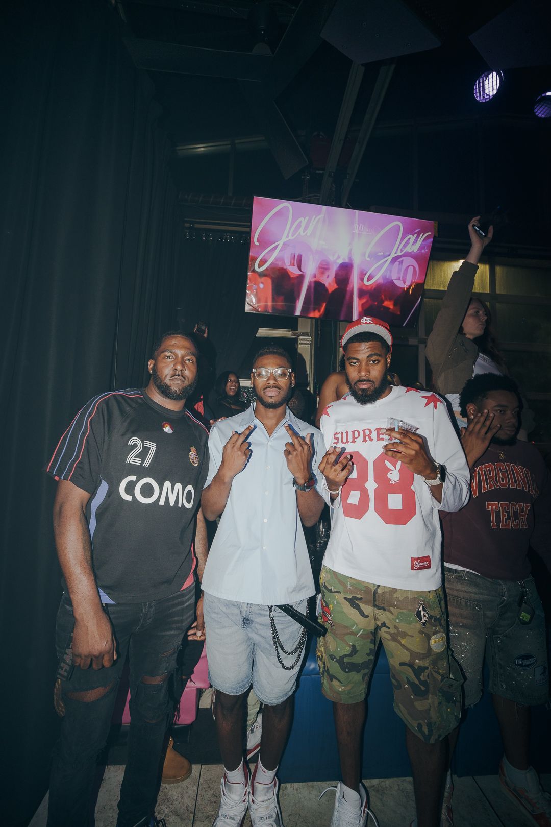 Three young men pose together at a nightclub event, one wearing a Como 27 jersey, another in white, and third in a Supreme shirt.