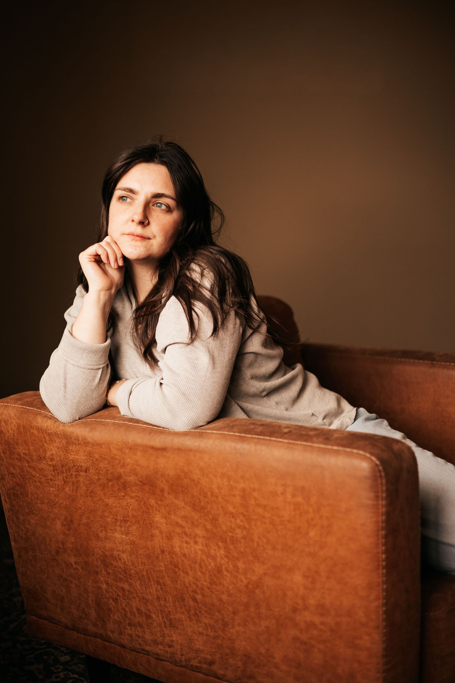 A contemplative young woman with dark hair rests her chin on her hand while leaning on a brown leather sofa |My Postpartum Depression Story