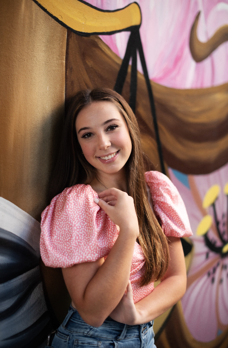 A person in a pink puff sleeve top and jeans poses against a colorful mural background with brown and pink tones.