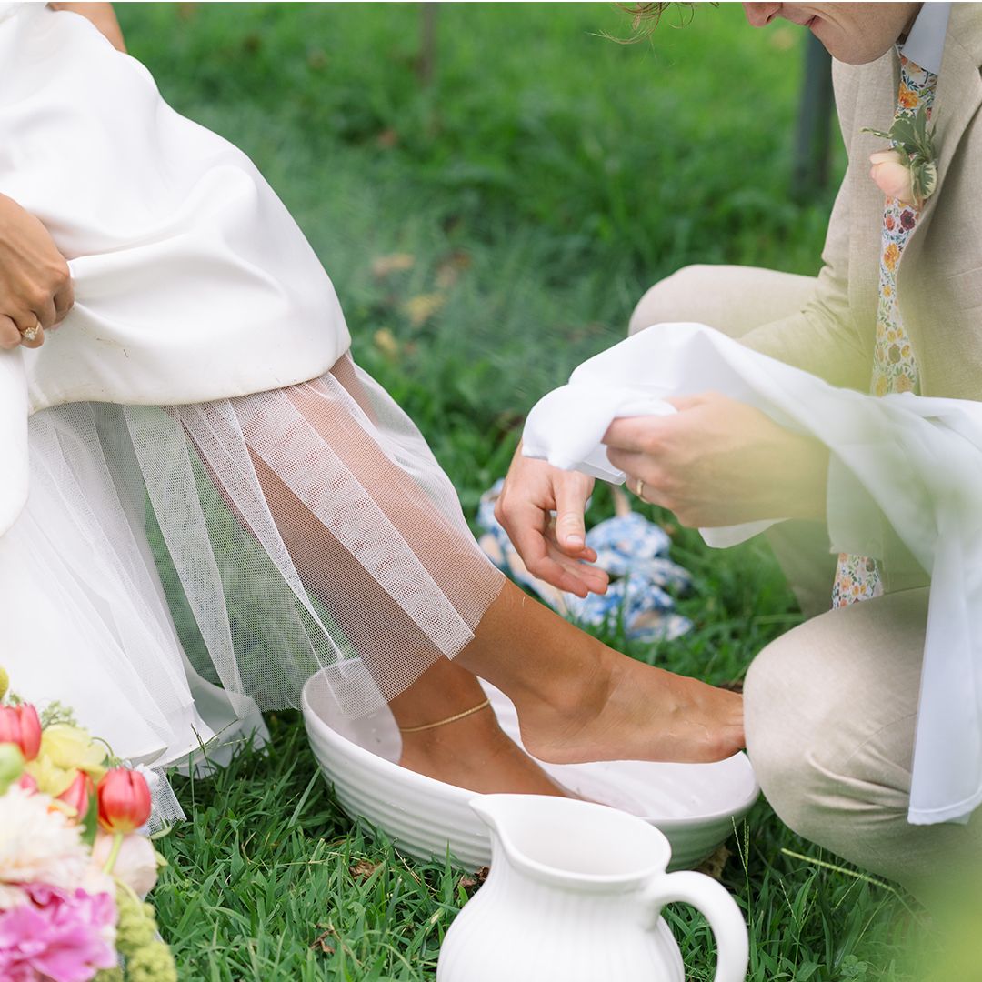 A child in a white dress sits on grass playing with a white ceramic teapot and teacups outdoors.