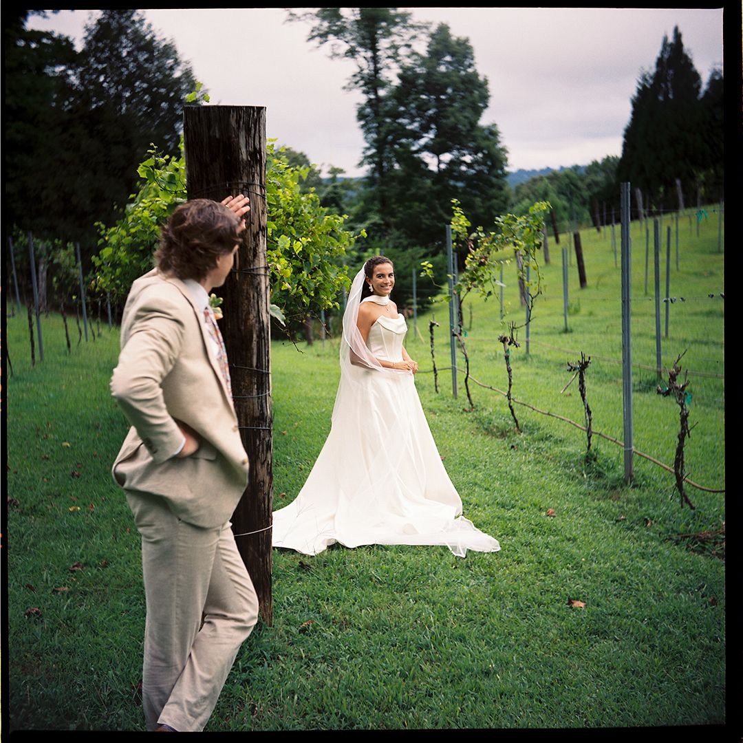 A couple in formal attire poses together in a scenic vineyard setting with rows of grapevines in the background.