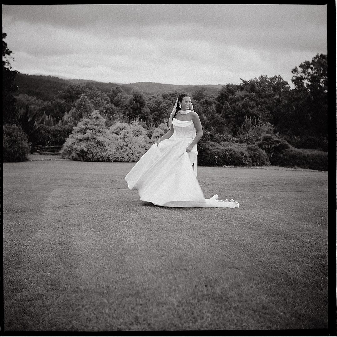A flowing white wedding dress billows in the wind on a hilltop lawn with mountain views in black and white photography.
