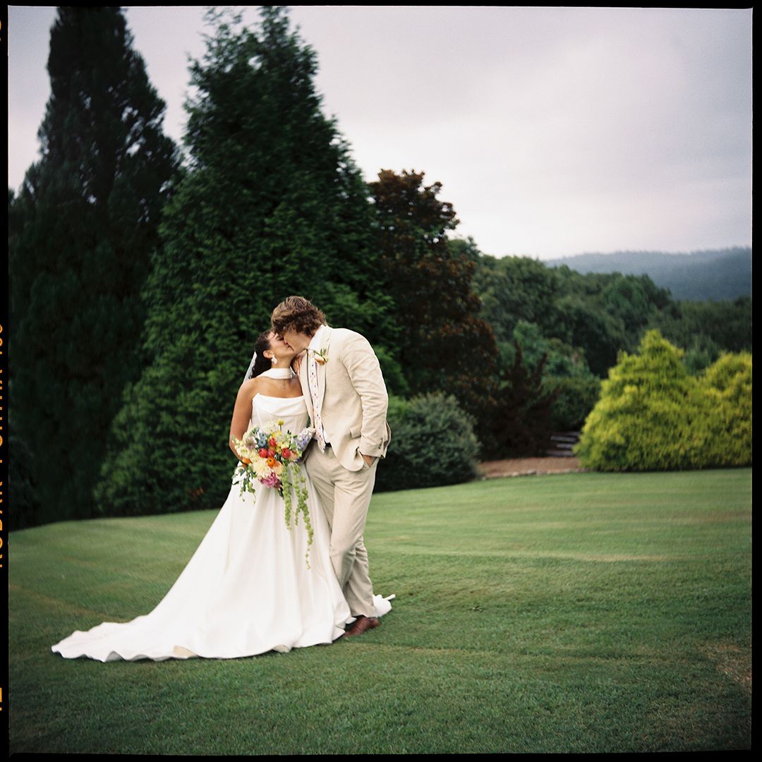 A couple in wedding attire shares an intimate moment on a scenic hilltop lawn surrounded by lush greenery.