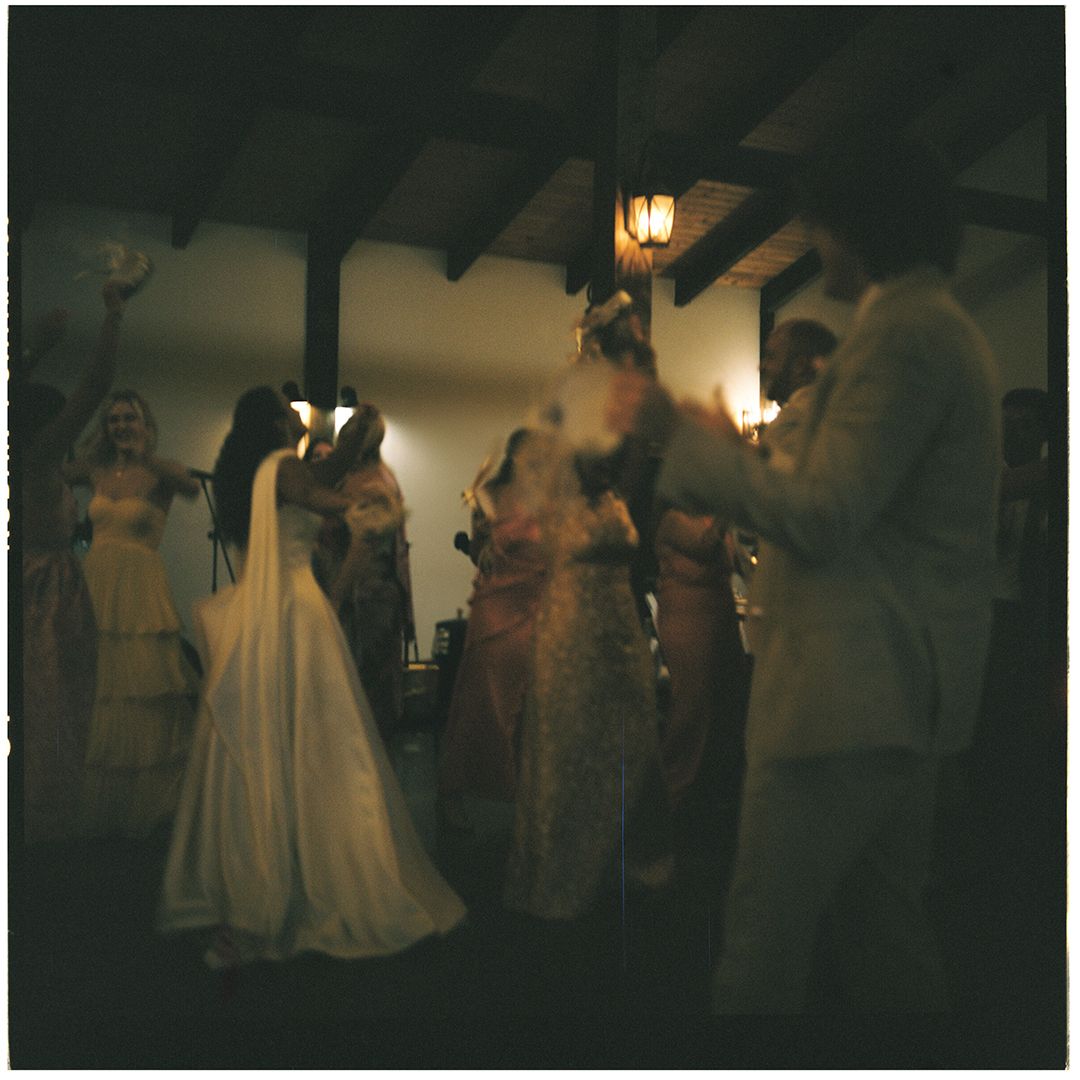 Guests in formal evening attire dance and socialize under warm lighting at an elegant indoor venue with rustic beams.
