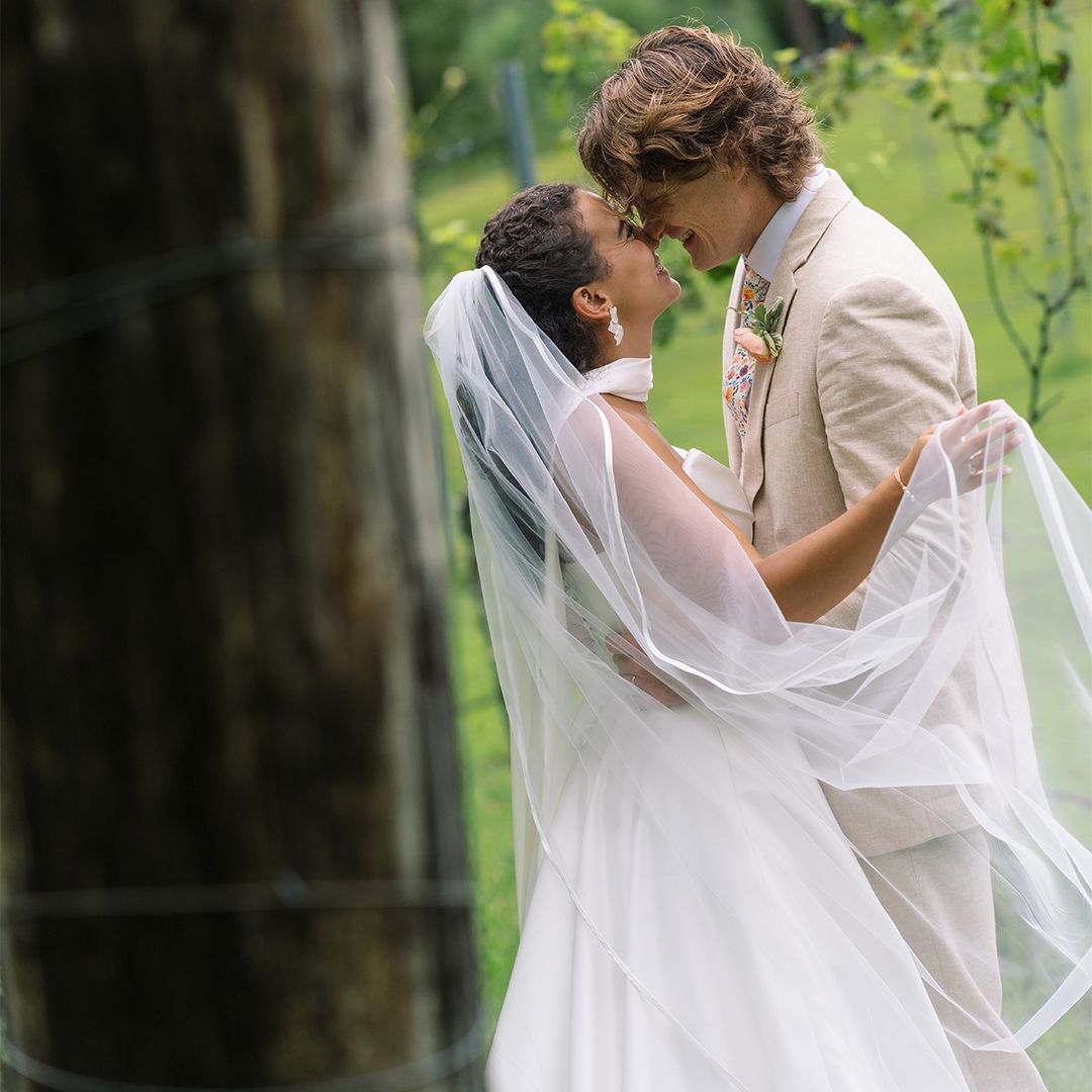 A romantic wedding couple embraces in a garden setting with flowing white veil creating an ethereal scene.