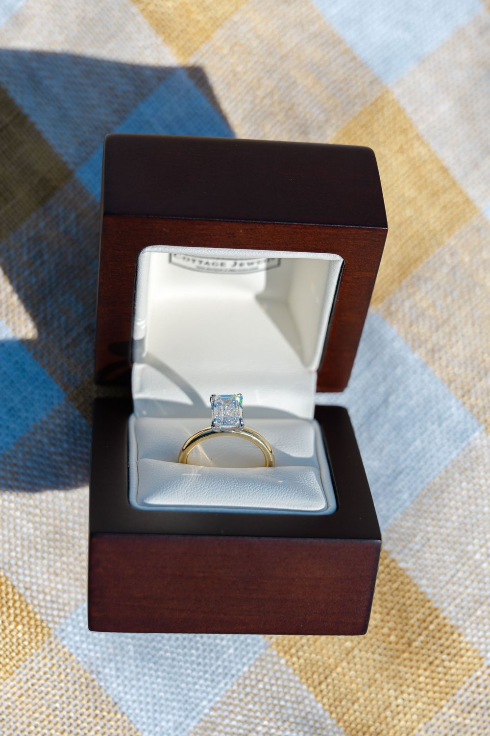 A gold engagement ring with a light blue stone sits in an elegant wooden ring box with white satin lining.
